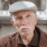 Elderly man in vintage jacket and cap sitting on a bench, reflecting a timeless style.